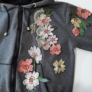 Puede incluir: Una sudadera con capucha gris con flores bordadas en tonos rosa, blanco y verde. Las flores están dispuestas en cascada en la parte delantera de la sudadera.