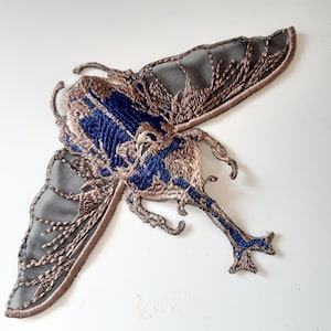 Peut inclure: Écusson papillon brodé avec des détails complexes. Le patch présente une combinaison de fils bleus, bruns et gris, créant un motif d'insecte détaillé. Les ailes sont en tissu gris transparent avec des broderies marron.