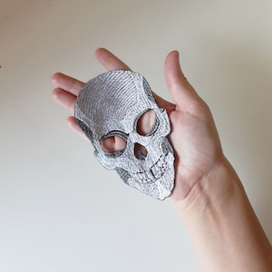 Könnte beinhalten: Ein silberner gestickter Totenkopf-Patch mit schwarzen Nähten. Der Schädel hat ein zahniges Grinsen und ist leicht zur Seite geneigt.