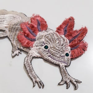 May include: Embroidered axolotl patch with pink and purple gills, white body, and green eyes. The axolotl is a type of salamander.
