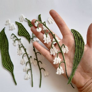 May include: White embroidered lily of the valley flowers with green stems and leaves. The flowers are arranged in a cluster and are attached to a hand.