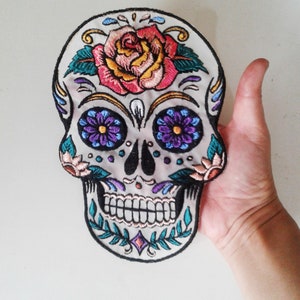 May include: Embroidered sugar skull patch with a white background, black outline, and colorful details. The skull has a pink rose, purple and blue flowers, and green leaves.