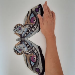 Könnte beinhalten: Gestickte Schmetterlingsflügel in Blau-, Grau- und Brauntönen mit lila Akzenten. Die Flügel sind symmetrisch und detailliert und weisen ein augenähnliches Design auf. Die Flügel sind vor einem weißen Hintergrund platziert.