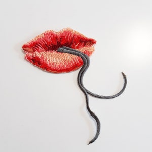 May include: Embroidered red lips with a black string attached. The lips are slightly parted and the string is coming out of the mouth.