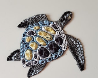 Pode incluir: Um remendo bordado de tartaruga marinha azul, preto e amarelo. O casco da tartaruga é detalhado com um padrão de círculos e espirais.