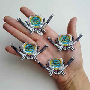 Könnte beinhalten: Vier gestickte Bügelbilder mit einem Spinnenmotiv in Blau, Gelb und Weiß. Die Spinne hat einen runden Körper mit einem detaillierten Design und acht Beinen.