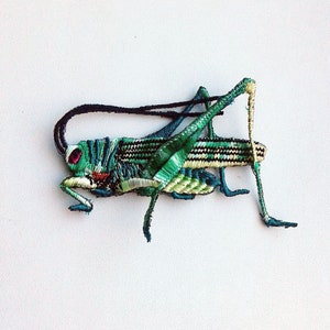 May include: An embroidered grasshopper made from thread, featuring green and black colours with red eyes. This is a decorative piece.