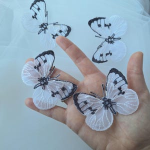 May include: White and black butterfly appliques with detailed wing patterns. The butterflies have delicate, lacy wings and dark accents. Four butterfly-shaped appliques are visible, with one held in a hand.