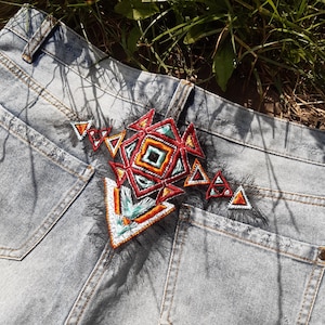 Op de afbeelding: Een paar blauwe denim jeans met een kleurrijk geborduurd patch op de achterzak. De patch heeft geometrische vormen in tinten rood, oranje, turquoise en wit.