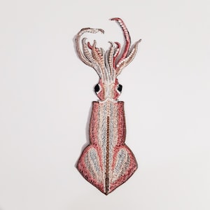 May include: Embroidered squid patch in shades of red, pink, and cream. The patch features detailed stitching, including the squid's tentacles, eyes, and body. This decorative patch is ideal for adding to clothing, bags, or other fabric items.