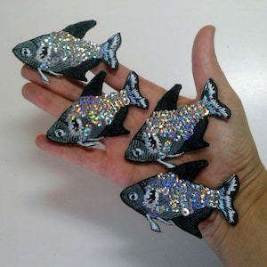 Op de afbeelding: Vier zilveren en zwarte visvormige patches met iriserende pailletten. De patches zijn geborduurd en hebben een gedetailleerd ontwerp.