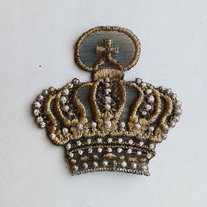 Puede incluir: Una corona bordada en oro y plata con una cruz en la parte superior. La corona es un parche o adorno decorativo.