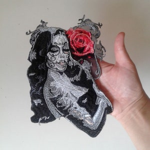 May include: Embroidered iron-on patch featuring a black and white sugar skull with a red rose. The skull has intricate details and a delicate floral design.