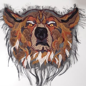 May include: Embroidered patch of a wolf's face with a fierce expression. The wolf's fur is depicted in shades of brown, orange, and gray, with a black nose and eyes. The patch has a frayed edge, giving it a rustic look.