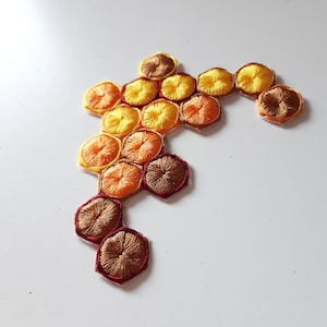 Op de afbeelding: Een honingraatvormige patch met geborduurde gele, oranje en bruine cirkels. De patch is gemaakt van stof en heeft een rode omtrek.