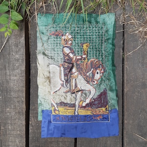 May include: A green and blue embroidered tapestry featuring a knight on horseback. The knight is wearing armor and holding a sword. The tapestry has the text "Knight's Crest" at the bottom.