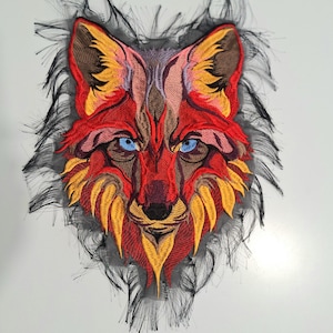 May include: Embroidered patch featuring a detailed wolf face. The design showcases a vibrant color palette of red, orange, yellow, and brown, with striking blue eyes. The wolf's face is surrounded by a black, textured outline, creating a bold and artistic statement.