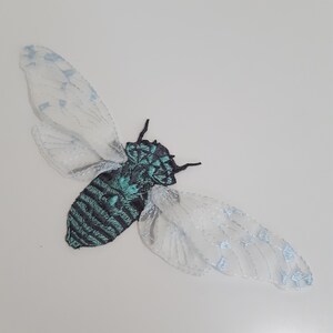 Pu&ograve; includere: Toppa ricamata a forma di cicala con corpo nero e turchese a righe. L'insetto ha ali delicate e trasparenti con dettagli azzurri. Questa toppa decorativa &egrave; ideale per aggiungere a vestiti o accessori.