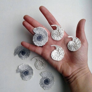 Puede incluir: Una colección de siete parches bordados a mano en forma de amonita, en blanco y gris. Los parches están dispuestos sobre una superficie blanca.