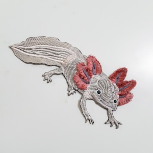 May include: Embroidered iron-on patch of an axolotl, a type of salamander, with a white body and pink feathery gills. The patch is perfect for adding a whimsical touch to clothing, bags, or other fabric items.