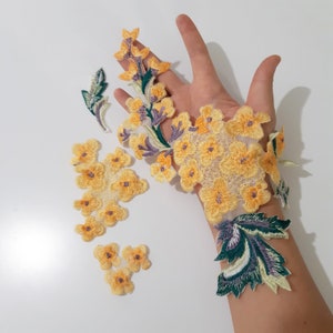 May include: Assortment of yellow floral appliques with green and purple accents. The appliques are arranged on a white surface and a hand.