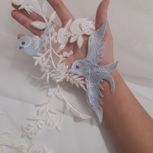 Può includere: Due uccelli ricamati blu con accenti floreali in pizzo bianco. Gli uccelli sono su uno sfondo bianco.