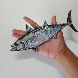May include: A silver and green embroidered iron-on patch of a fish with iridescent scales.