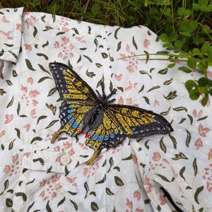 Könnte beinhalten: Ein gelber und schwarzer Schmetterling mit blauen Akzenten ist auf einem weißen Stoff mit Blumenmuster gestickt.
