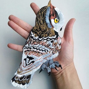 May include: Embroidered iron-on patch of a brown, white, and gray owl with yellow eyes. The owl is facing to the right and has its wings spread out.