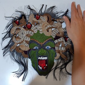 May include: A close-up of a green and brown embroidered patch depicting Medusa, a mythical Greek figure with snakes for hair. The patch is on a white background.