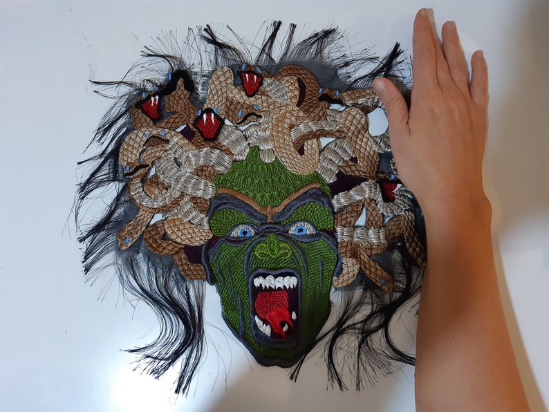 May include: A close-up of a green and brown embroidered patch depicting Medusa, a mythical Greek figure with snakes for hair. The patch is on a white background.