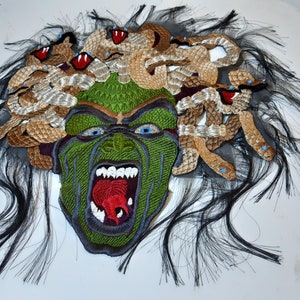 May include: A green and brown embroidered patch depicting a monstrous face with snakes for hair. The face is open-mouthed, revealing sharp teeth and a red tongue. The snakes are brown and gold, with blue eyes.
