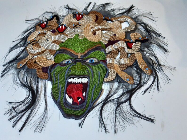 May include: A green and brown embroidered patch depicting a monstrous face with snakes for hair. The face is open-mouthed, revealing sharp teeth and a red tongue. The snakes are brown and gold, with blue eyes.