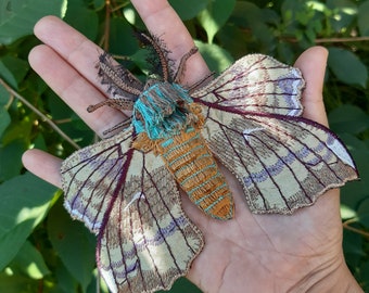 May include: A hand-stitched embroidered moth with brown, beige, and purple wings. The moth has a turquoise and orange body.
