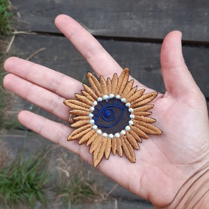 May include: A gold embroidered patch featuring a blue eye at its centre, surrounded by white beads. The eye is encircled by a dark blue circle and has a white pupil. The patch is shaped like a flower with numerous petals.