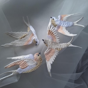 May include: Embroidered swallows in flight, featuring detailed stitching in white, tan, and pale blue. The birds have outstretched wings and are arranged against a soft, neutral background, creating a delicate and artistic design.