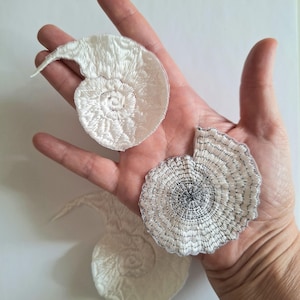 Puede incluir: Tres parches de conchas bordadas en blanco con diseños en espiral. Los parches están en varias etapas de finalización, con uno completamente formado y dos parcialmente terminados. Los parches están hechos de una tela suave y texturizada.