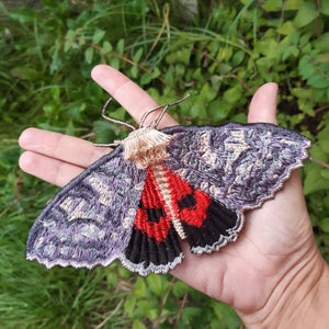Puede incluir: Una polilla bordada a mano con un cuerpo estampado en morado y gris y alas rojas y negras.