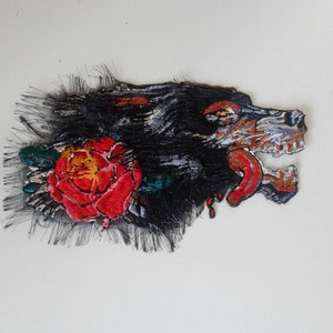 Op de afbeelding: Zwarte en witte geborduurde patch van een grommende wolf met een rode roos en groene bladeren. De tong van de wolf steekt uit en er komen rode bloeddruppels uit zijn bek.
