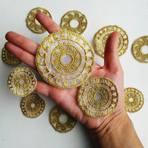 May include: A hand holding eight gold and white circular lace appliques. The appliques have a delicate, intricate design.