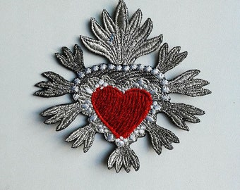 Könnte beinhalten: Ein silberner gestickter Aufnäher mit einem roten Herzen in der Mitte. Das Herz ist von einem silbernen Heiligenschein mit weißen Perlen umgeben. Der Aufnäher hat ein Sternburst-Design mit mehreren Punkten.