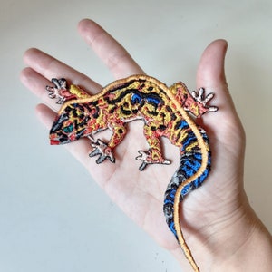 May include: Embroidered lizard patch in shades of orange, yellow, blue, and black. The patch features intricate stitching details, including the lizard's legs, tail, and head. The lizard is approximately 15 cm long.
