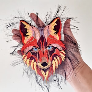 Puede incluir: Parche bordado de cabeza de zorro rojo, naranja y amarillo con ojos azules. El parche está sobre una tela de malla negra con bordes deshilachados.