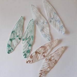 Può includere: Cinque ali di tessuto ricamate in vari colori, tra cui azzurro, verde e oro. Le ali sono delicate e traslucide, con motivi intricati.