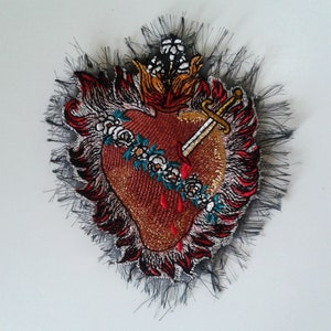 Peut inclure: Un cœur rouge brodé avec une épée dorée qui le transperce, entouré de fleurs blanches et de flammes. Le cœur est sur un fond de dentelle noire.