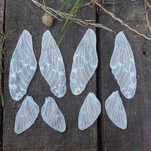 Może przedstawiać: Kolekcja ośmiu delikatnych, jasnoszarych skrzydeł wróżki z tkaniny z misternymi białymi i jasnoniebieskimi haftowanymi detalami. Skrzydła ułożone są na zwietrzałej drewnianej powierzchni, prezentując ich szczegółowy design i eteryczną jakość.