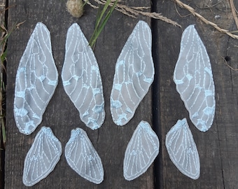 Puede incluir: Una colección de ocho delicadas alas de hadas de tela gris claro con intrincados detalles bordados en blanco y azul claro. Las alas están dispuestas sobre una superficie de madera envejecida, mostrando su diseño detallado y calidad etérea.