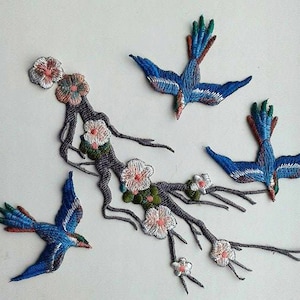 May include: Embroidered iron-on patches featuring a floral branch with pink and white flowers and three blue birds. The birds have green, blue, and white accents.