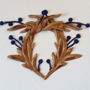 May include: A gold-colored fabric applique in the shape of a wreath with blue berries. The wreath is made of leaves and branches.