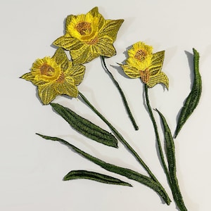 May include: Three yellow embroidered daffodil flowers with green stems and leaves. The flowers are arranged in a bouquet.